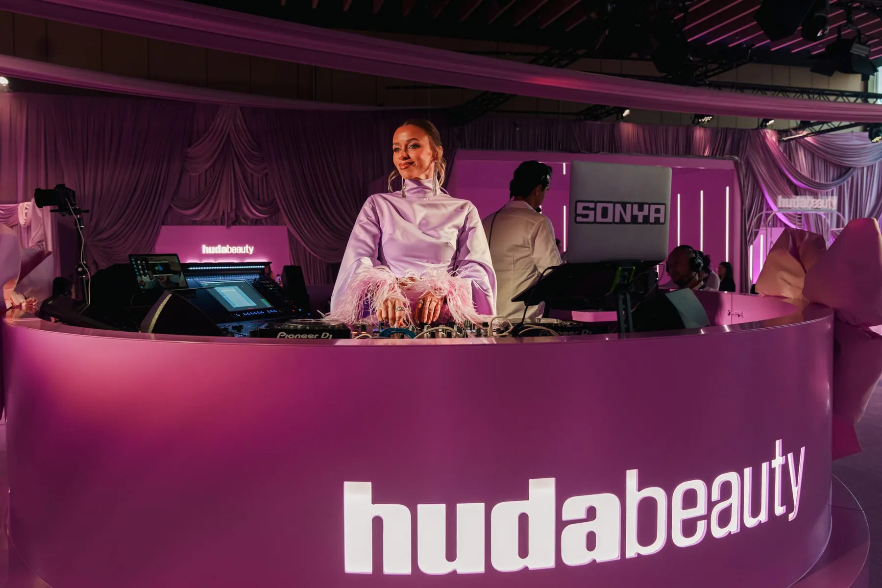 The Huda Beauty UBE launch in the UAE, a magenta-lit stage with a DJ booth carrying the hudabeauty wordmark